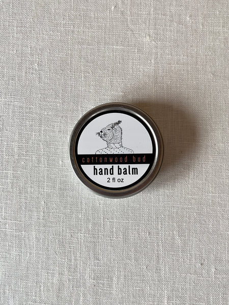 A round aluminum container with a white label. Theres a fuzzy animal in a polkadot sweater on the label. There's black and orange text under the animal that reads "Cottonwood Bud hand balm 2 fl oz"