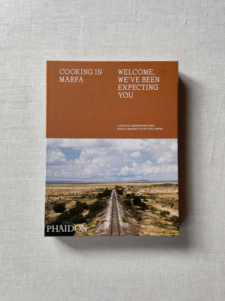Cover of the book "Cooking In Marfa" by Virginia Lebermann and Rocky Barnette of The Capri. The cover has a textured orange top and a desert scene below it. Additional text reads "Cooking in Marfa: Welcome We've Been Expecting You." 