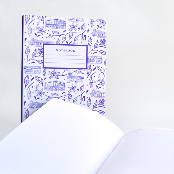 Notebook opened to show blank pages with a closed notebook behind it. The closed notebook features purple artwork of famous buildings in Albuquerque, NM separated by flowers and leaves. In the middle of the art there is a rectangle with the lettering "notebook" and three lines under the lettering to label the notebook.