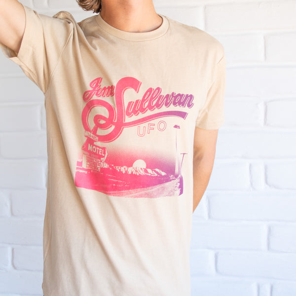 Cream shirt showing a desert landscape with a 60's motel in the corner. at the top is pink stylized lettering reading "Jim Sullivan UFO."