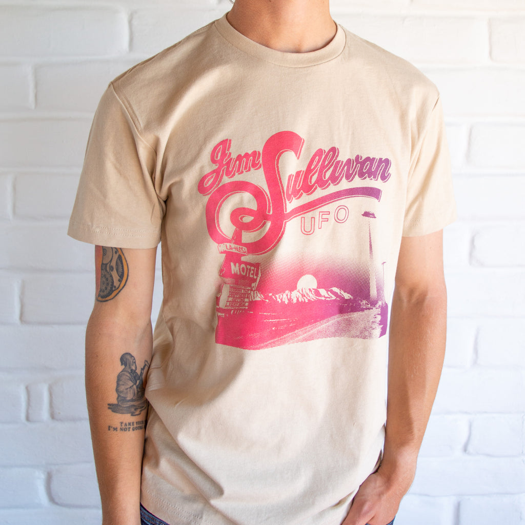Cream shirt showing a desert landscape with a 60's motel in the corner. at the top is pink stylized lettering reading "Jim Sullivan UFO."