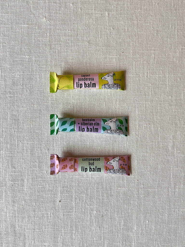 Three cardboard tubes of lip balm on a white backdrop. From top To Bottom: green and pink "sweet ponderosa" lip balm, green and purple "beebalm + siberian elm" lip balm, and Pink and orange "cottonwood bud" lip balm.