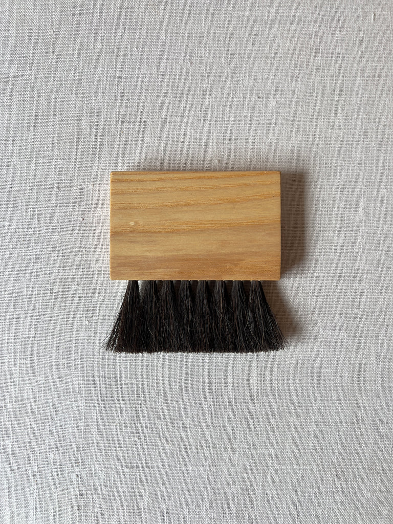 Square handheld broom with light wood and black horsehair bristles.