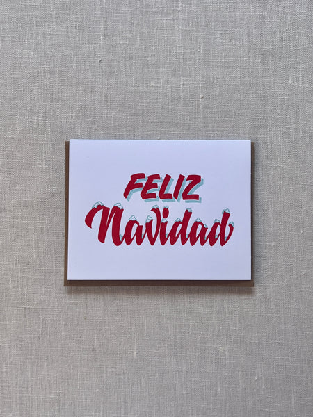 White card with red lettering reading "Feliz Navidad." the lettering looks as if snow has fallen on them.