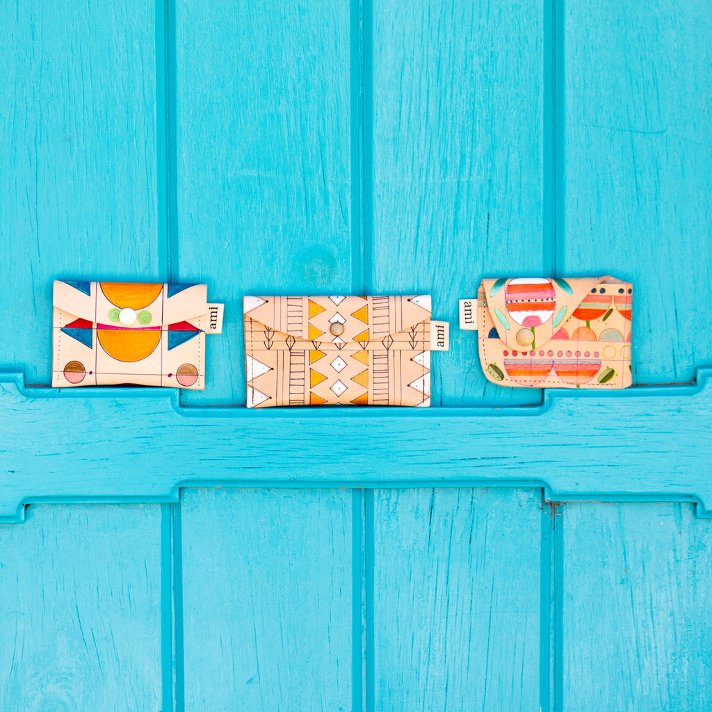 Three tan leather pouches laying against a bright blue painted wood. The pouches lay left to right in a horizontal manner, and have shapes of varying size and color painted on them.