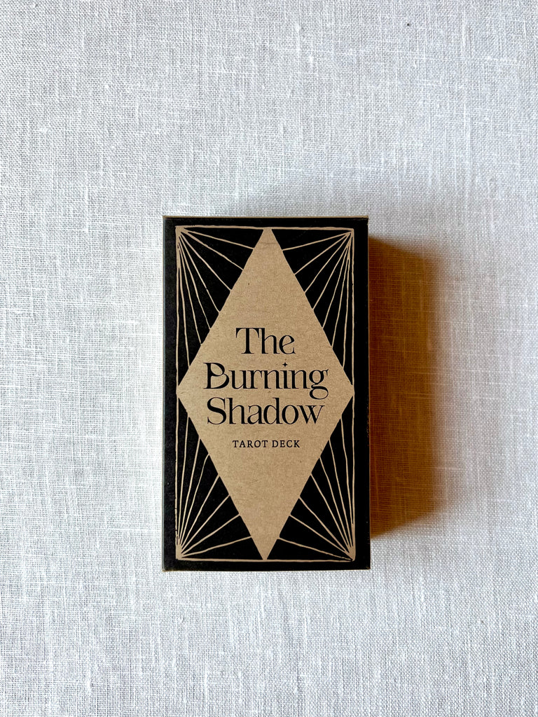 Front of a cardboard container for a tarot card deck. The deck has a black and tan design with a large tan diamond in the middle. In the middle of the diamond there is text reading "The Burning Shadow Tarot Deck."