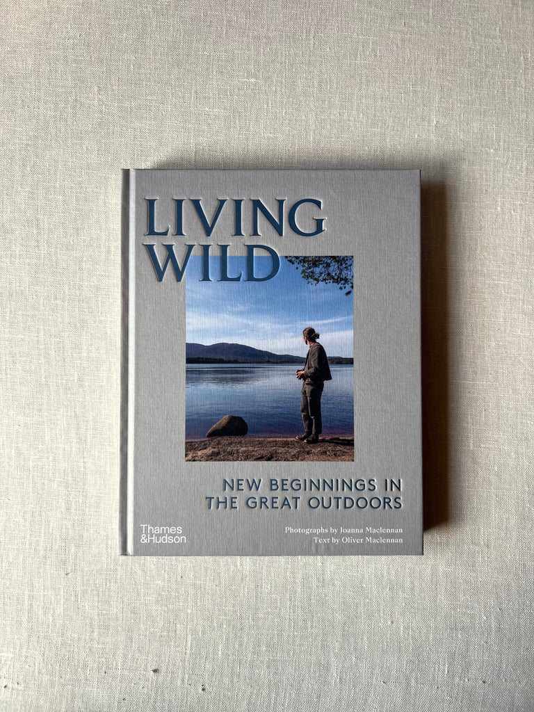 Grey cover of the book "Living Wild: New Beginnings In The Great Outdoors"  in the middle is a picture of a man looking out onto a river with mountains behind it