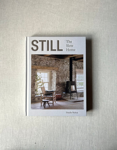 A cover of the book "Still: The Slow Home" by Natalie Walton. The cover is white with a photo of a living room in a cabin with a wooden stove sitting next to a wooden chair and stool, surrounded by two large windows with lots of light entering the room.