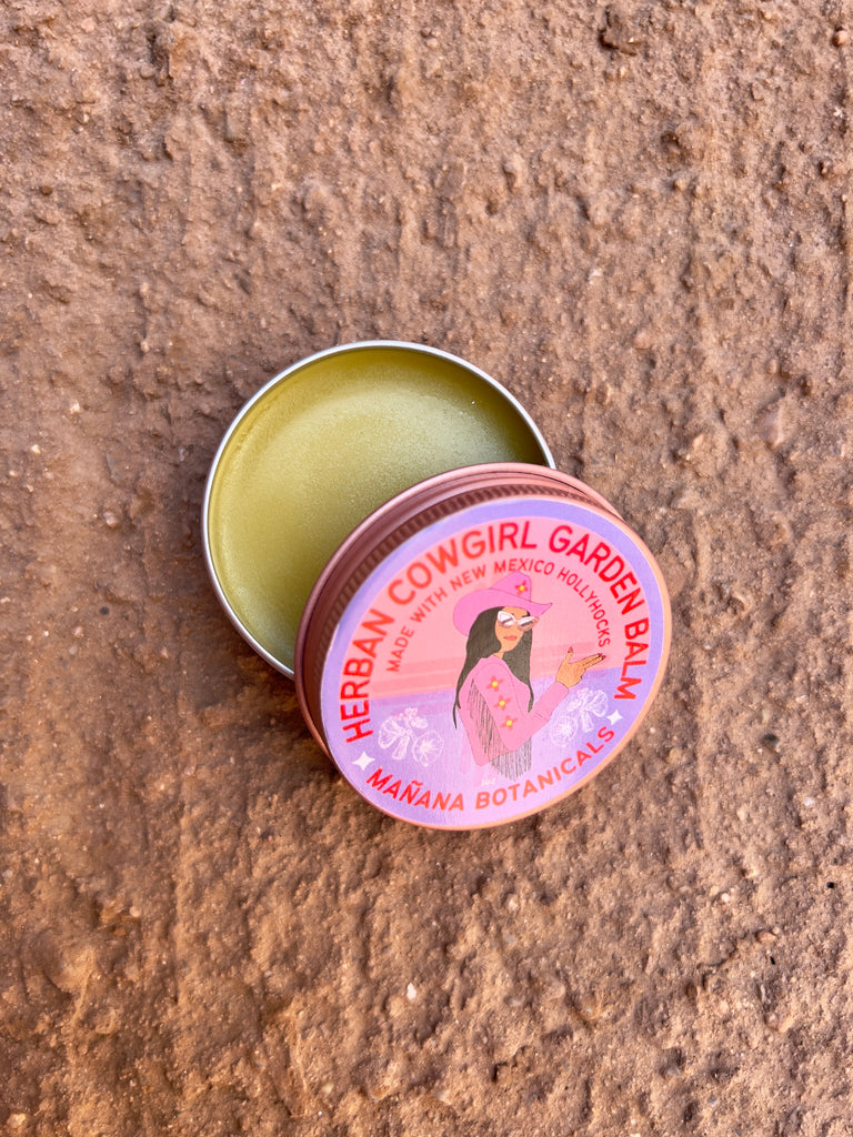 Circular tin can with red text circling a cowgirl in pink clothing. The text reads "Herban Cowgirl Garden Balm. Made with New Mexico HollyHocks. Mañana Botanicals." the tin is open to show the light yellow balm inside.
