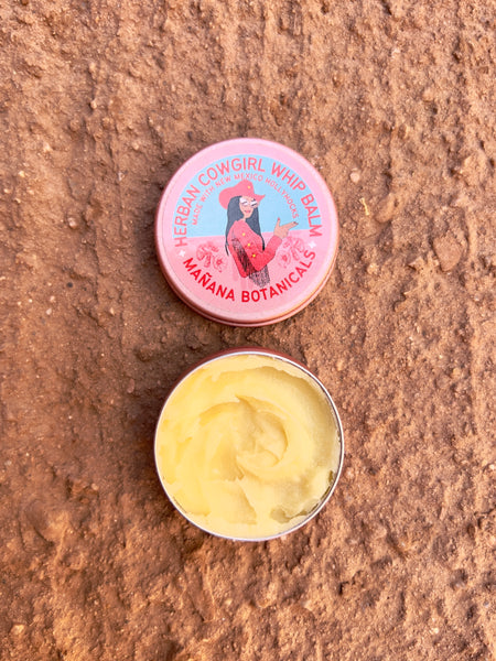 Circular tin can with red text circling a cowgirl in pink clothing. The text reads "Herban Cowgirl Garden Balm. Made with New Mexico HollyHocks. Mañana Botanicals." The tin is opened to show the light yellow whipped balm inside