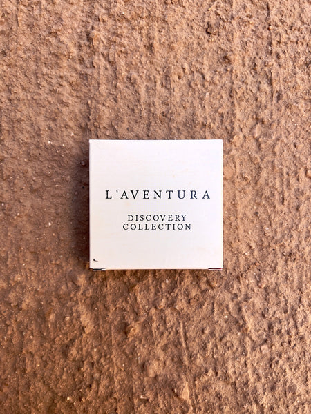 Small, square, white paper box. The box has black text reading "L'aventura Discovery Collection."