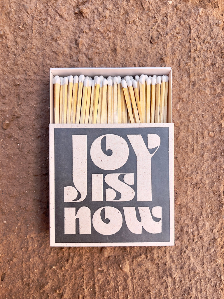 Square Matchbox with tan text readign "Joy is now" on a black base. The box is open to show the long, white tipped matches inside.