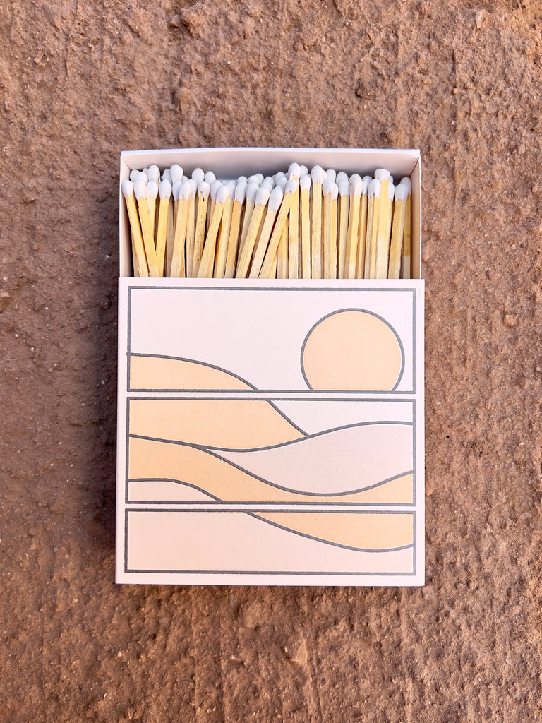 Square match box with a pink and orange sunset desert scene. the box is open to show the long, white tipped matches inside.