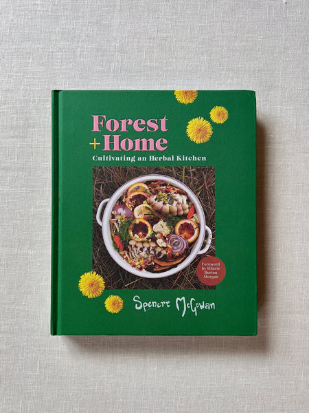 Rich green cover of the book "Forest + Home" by Spence McGowan.There is an image of a cooking pot sitting in grass with loads of herbs, mushrooms, fruits and vegetables placed in it. Additional text reads "Forest + Home: Cultivating an Herbal Kitchen."
