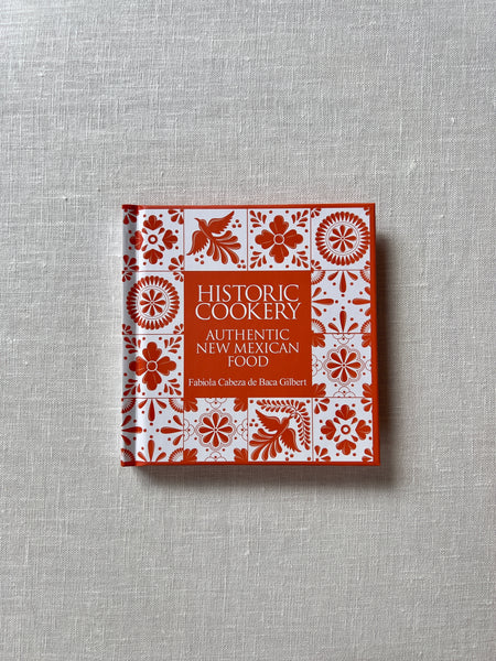 Cover of the small square book "Historic Cookery, Authentic New Mexican Food" by Fabiola Cabeza de Baca Gilbert. The cover is orange and replicates spanish tiles.