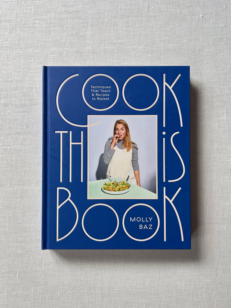 Rich blue cover of "Cook this book" by Molly Baz. A picture of a woman taking a bite of a salad that sits in front of her. Text reads Cook This Book: Techniques that Teach & Recipes to Repeat.