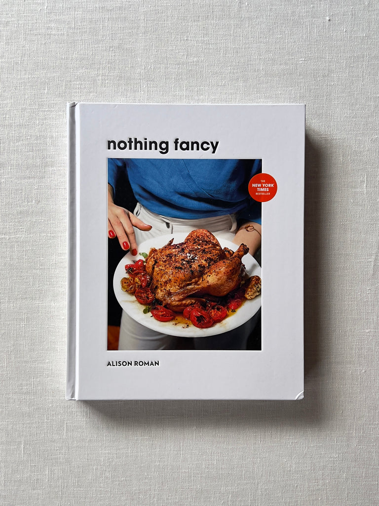 White cover of the book "Nothing Fancy" by Allison Roman. the middle of the the cover sows someone holding spiced chicken.