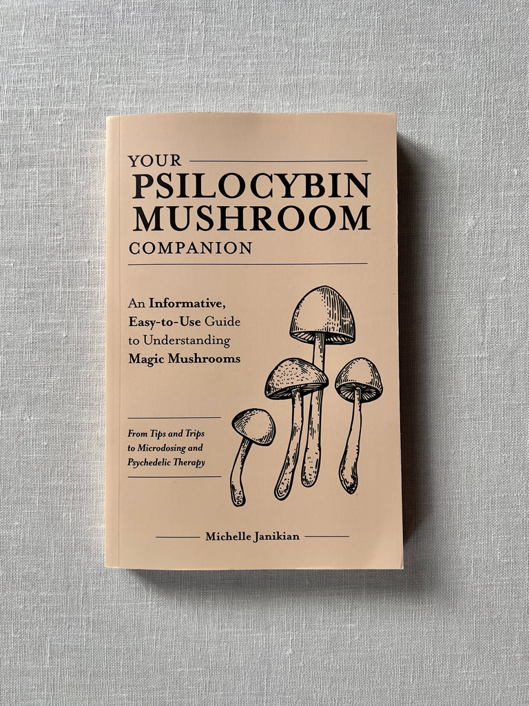 A cover of the book "Your Psilocybin Mushroom Companion" by Michelle Janikian. The cover has text and an illustration of a group of mushrooms. Additional text reads "An Informative Easy-to-Use Guide to Understanding Magic Mushrooms. From Tips and Trips to Microdosing and Psychedelic Therapy."