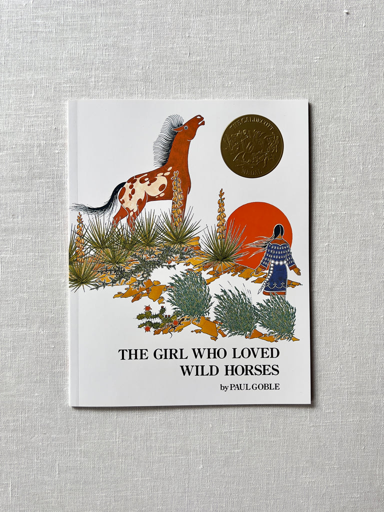 A cover of the book "The Girl Who Loved Wild Horses" by Paul Globe. The cover is white with a very colorful illustration of an Indigenous girl who stands facing the sun and a wild horse, surrounded by wild desert plants. 