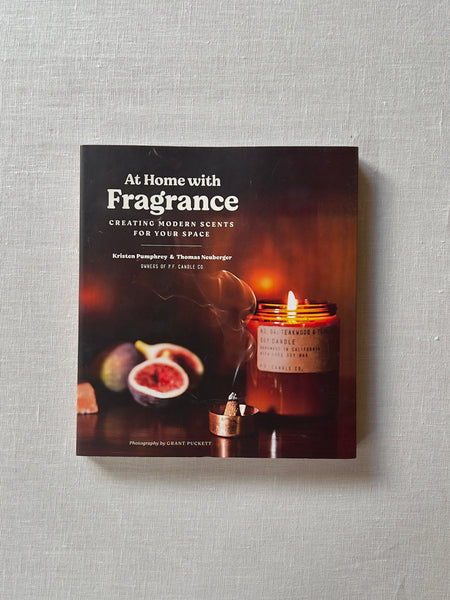 Book with various shades of dark orange and brown. In the rightmost corner there is lettering reading "At home with fragrance" "Creating modern scents for your space" "Kristen Pumphrey & Thomas Neuberger owners of P.F. Candle Co." Diagonal of the writing is a lit candle in amber glass with a burning cone incense in front. even farther back to the left and a bit out of focus are cut figs and pink salt. The bottom of the cover reads "Photography by Grant Puckett"
