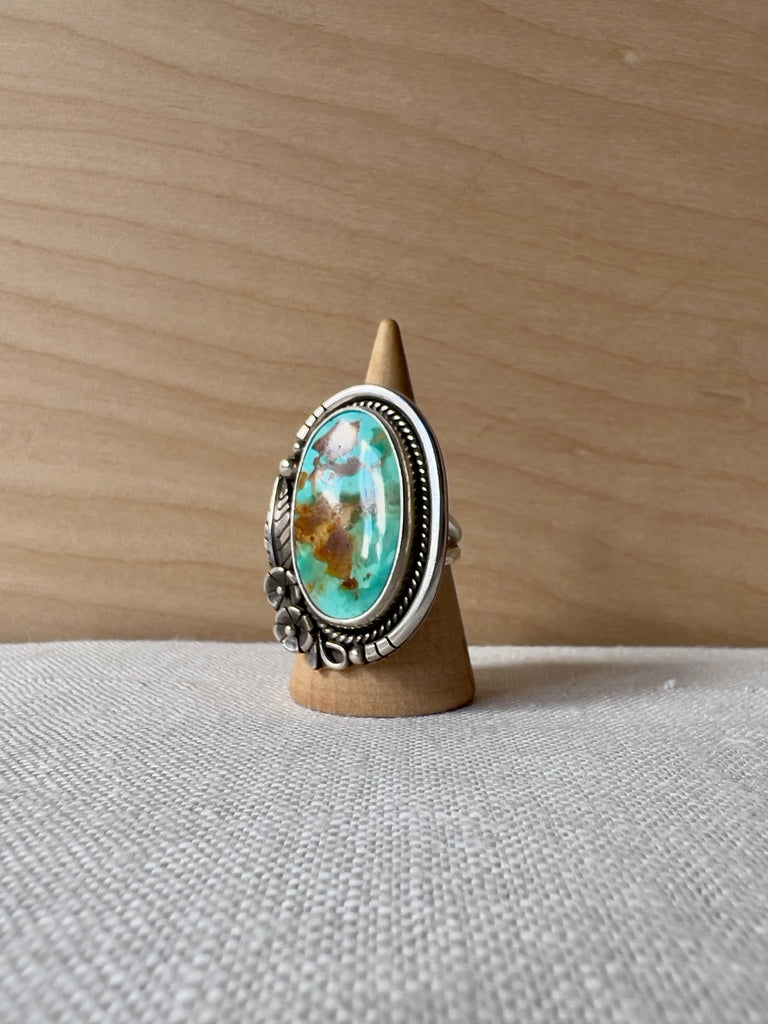 Oval turquoise ring with a rounded sterling silver casting with feathers and flowers at one corner
