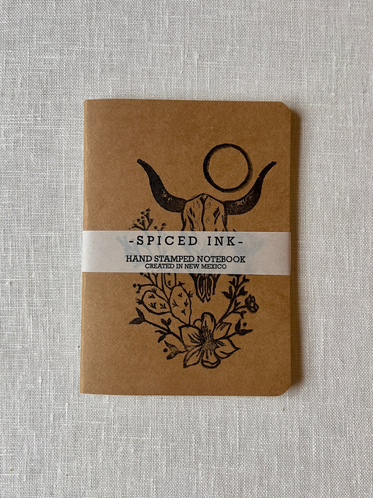 Small brown paper notebook with a horned cow skull, cactus, circular sun,  and vined flowers in black in on the front. there is a white band around the notebook with black text reading "Spiced Ink, hand stamped notebook created in new mexico."
