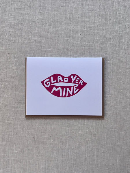 White card with red lips in the middle. the lips have white text on the top and bottom reading "Glad yer mine."