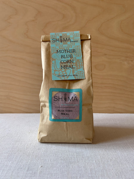 Brown paper bag sealed with a blue and brown label at the top reading "Shima Mother Blue Corn Meal"