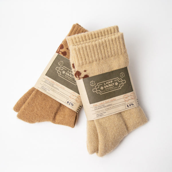 Two pairs of tan socks, one darker than the other.  The lighter pair is resting atop the darker pair at a 45 degree angle. Both pairs have a tan and army green paper label with lettering reading "Land object knitwear fine camel wear socks" "softer than wool, more durable than cashmere" "indigenous designed and manufactured" "70% camel hair, 30% nylon."