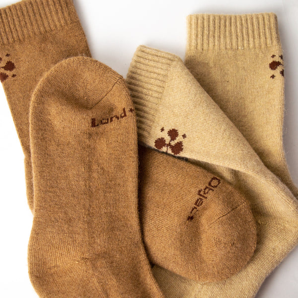 A pair of dark tan socks and a pair of light tan socks on top of one another. The cuffs of the socks are ribbed and there are dark brown geometric shapes woven in at the back. The words "land" and "object are seen on the tops of the socks.