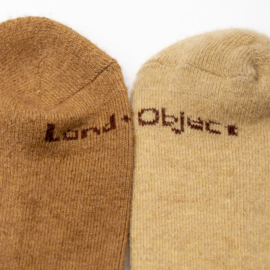 a closeup of one dark tan sock and one light tan sock laying next to each other showcasing the words "land object" woven into the bottom.