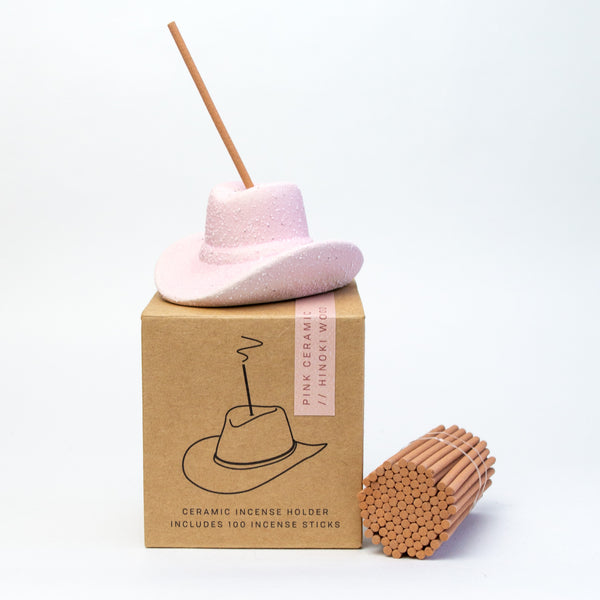 Pink ceramic cowboy hat sitting on top of a small cardboard box. The box has a drawing of the same hat that is atop it and black lettering that reads "Ceramic incense holder includes 100 incense sticks. A bundle of small incense sticks is laying next to the box and hat.