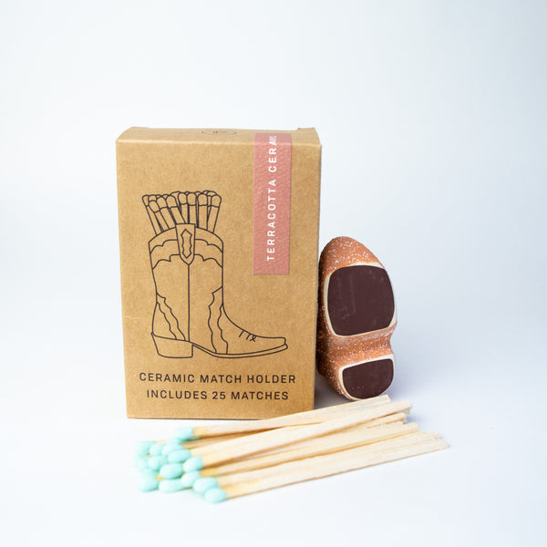Small tan cardboard box with art of a cowboy boot full of matches and black text under it reading "ceramic match holder includes 25 matches." Next to the box is a small ceramic boot, laid to show its sole. There are matches with blue tips in front of the box and small boot.
