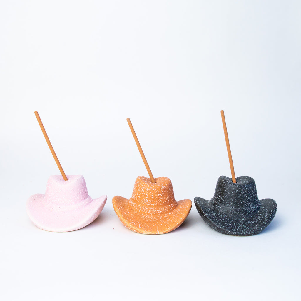 Three small ceramic cowboy hats sitting next to one another, all with small incense sticks coming out of the tops of each. From left to right pink, terracotta, and black.
