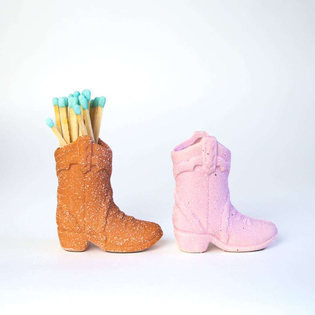 Two small ceramic cowboy boots, one is terracotta (left) and one is pink (right). The terracotta boot is filled with matches that have a white tip.