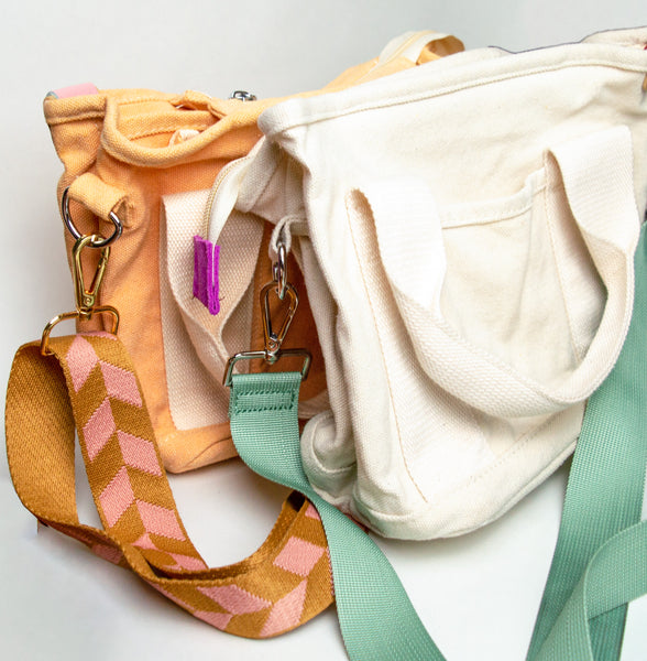 Two medium canvas bags, one in front of the other. From back to front: light orange canvas bag with white handles attached to the body and a checkered orange and pink cross-body strap hooked to the bag with a brass clasp and silver loop. The front most bag is white with white handles attached to the body, one small front pockets and a turquoise colored cross-body strap  connected my a silver clasp and loop. 