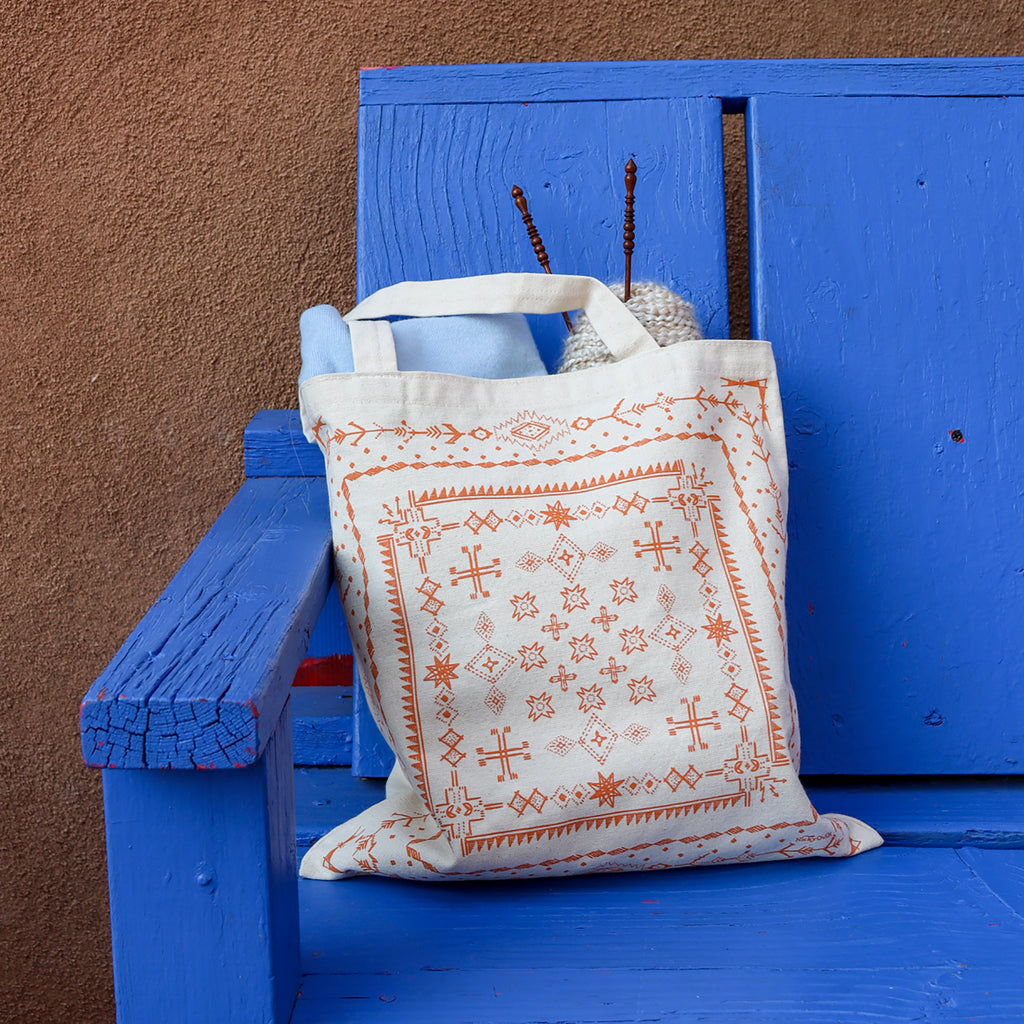 Orange southwestern patterns printed on a white canvas tote. Filled with various items. The tote is sitting on a blue bench.