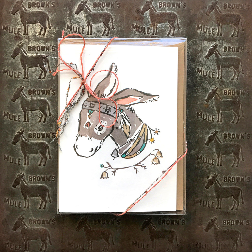 A grey donkey with a white mouth and nose wearing a silver headpiece, a silver bell and necklaces on a card with a white background.