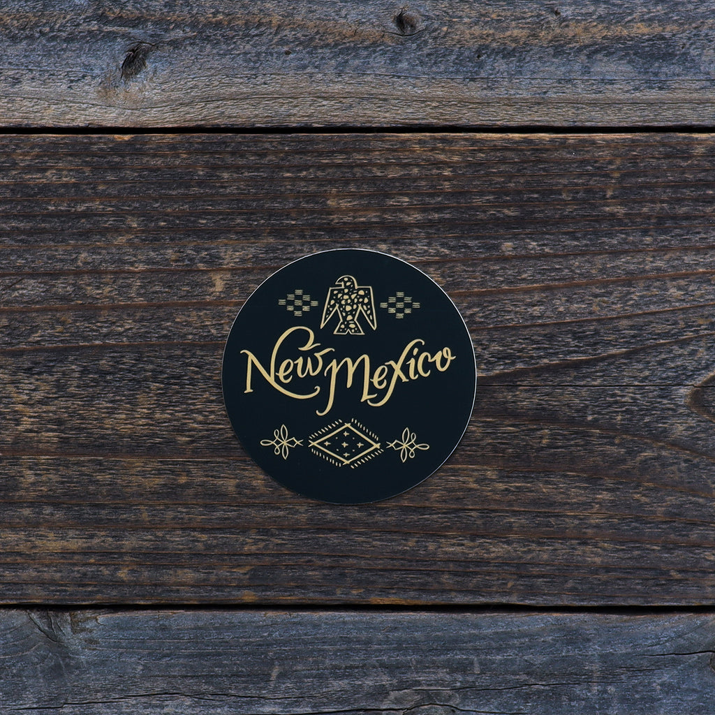 Circular sticker with a black background and yellow text reading "New Mexico." Theres southwestern designs at the top and bottom of the sticker.
