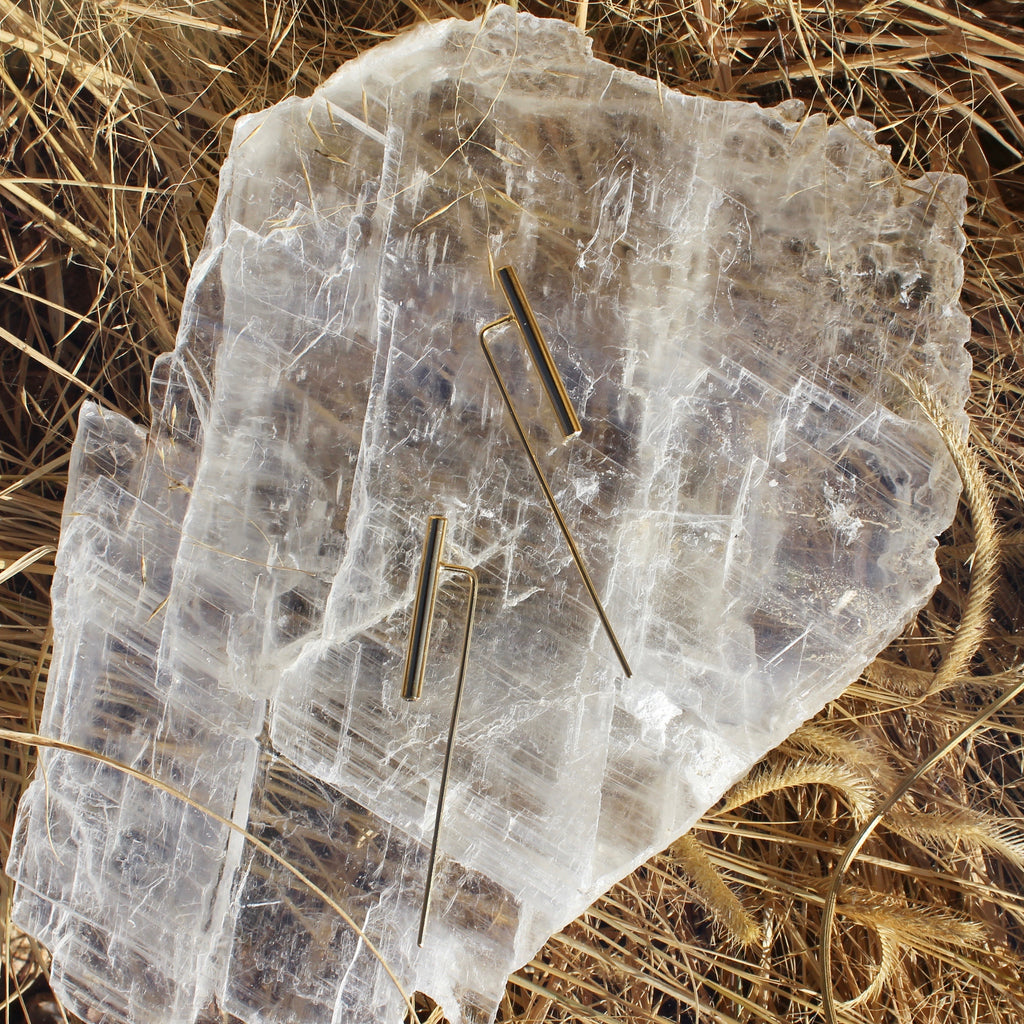 Golden long earrings with very long finding. Parallel to the finding is a vertical barbell about half its size. The earrings are laying atop a large quartz slab.