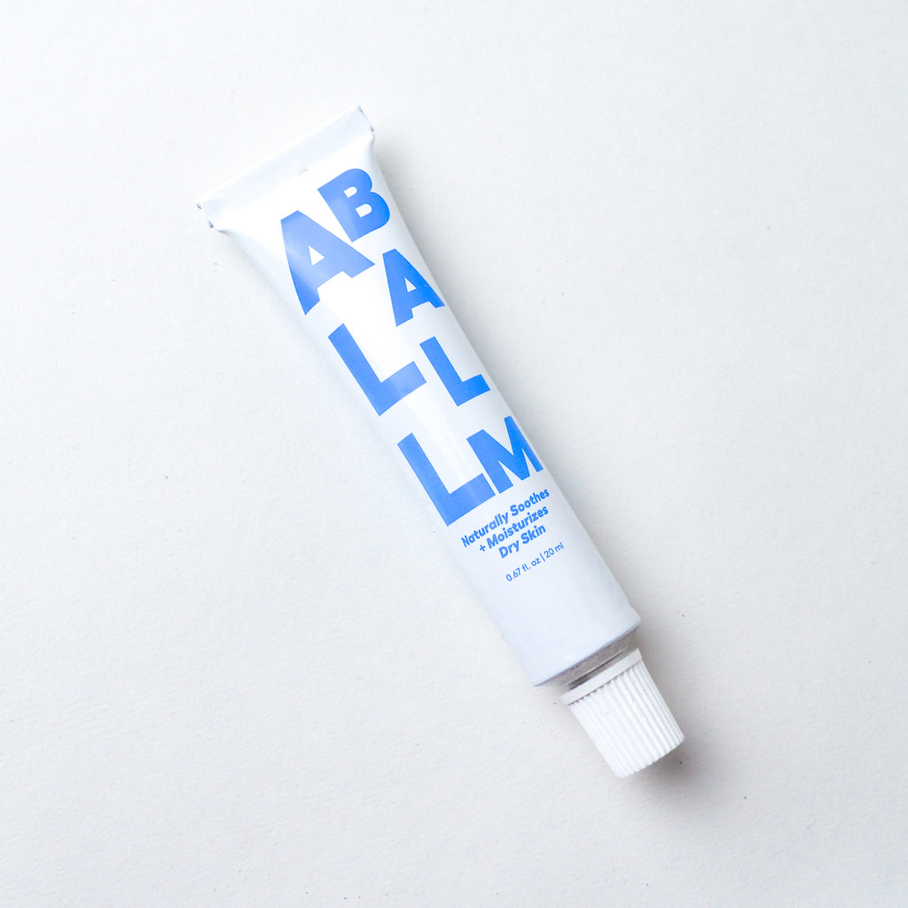 White aluminum tube resembling the style of a toothpaste container. The tube has blue stylized lettering reading "All Balm" "Naturally soothes + moisturizes dry skin" "0.67 fl. oz | 20 ml." The lettering is trailing vertically down the tube.