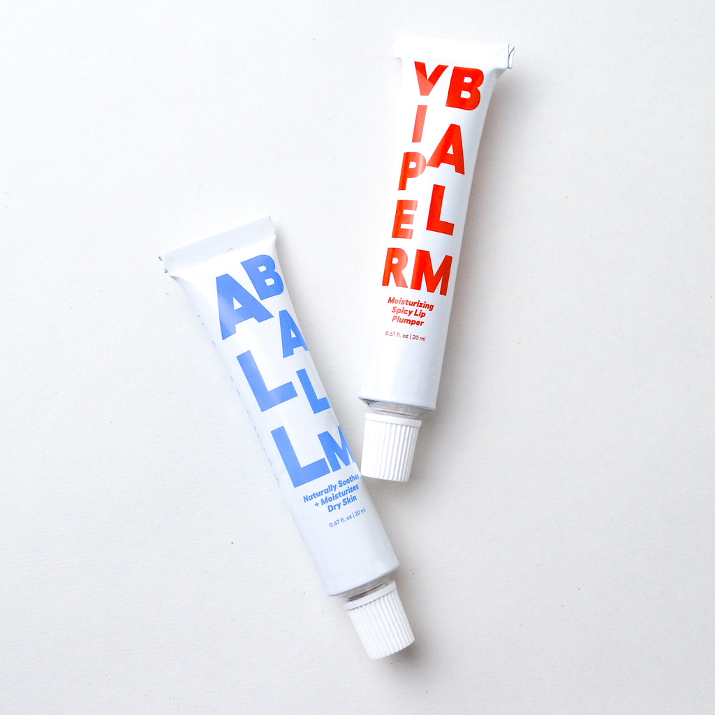 Two white aluminum tubes laying asymmetrically. The leftmost tube reads "All Balm" "Naturally soothes + moisturizes dry skin" "0.67 fl. oz | 20 ml" in blue lettering. The rightmost tube reads  "Viper balm" "Moisturizing spicy lip plumper" "0.67 fl oz | 20 ml" in red lettering.