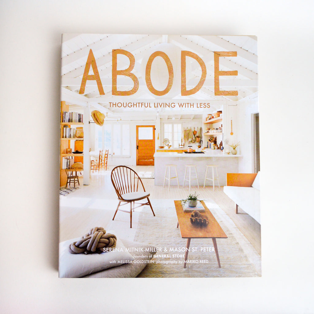 A Book which reads "Abode Thoughtful Living with Less" at the top. Pictured behind and below the lettering a picture of a home scene, from the living room looking into the kitchen. The home scene is in neutral colors like white and brown. The bottom of the book reads "Serena Mitnik-Miller & Mason ST. Peter Founders of General Store with Melissa Goldstein photography by Mariko Reed" in white text.