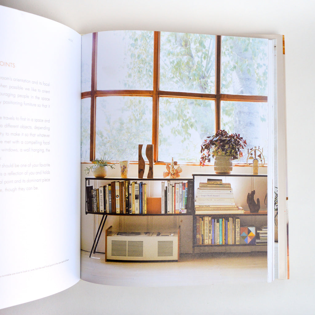 Page of "Abode" book showcasing the decoration of a simple media console in front of a large window. Media console is filled with books of varying size and color. On top of the console are various vases and modern art pieces. Below the console is a large record player. The colors of the photo are of a neutral color palette. Style of the items would be considered mid century modern.