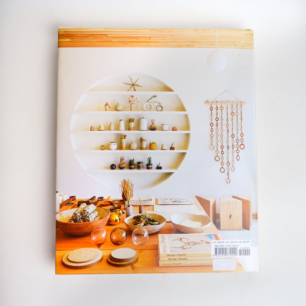 The back of the book "Abode" which is showing a scene of a highly decorated room. There is a table and shelf both full of various items set with intention. The room is of neutral colors.