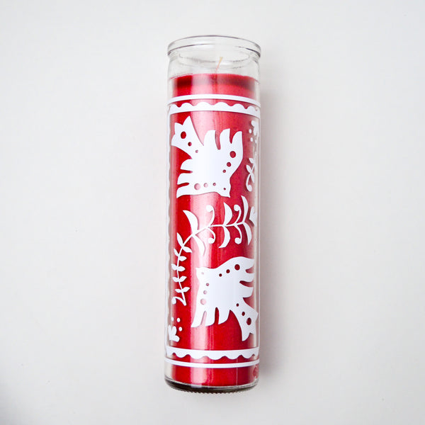 Tall red candle in a glass vessel with white designs of doves and branches.