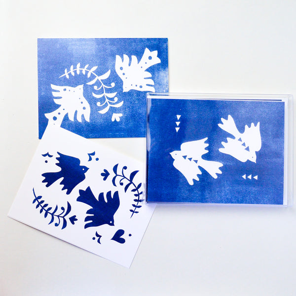 Three cards, one with a white background, two with a dark blue background. On top of each of the cards are two doves and olive leaves. there's variation in each of the cards with different shapes on or around the doves.