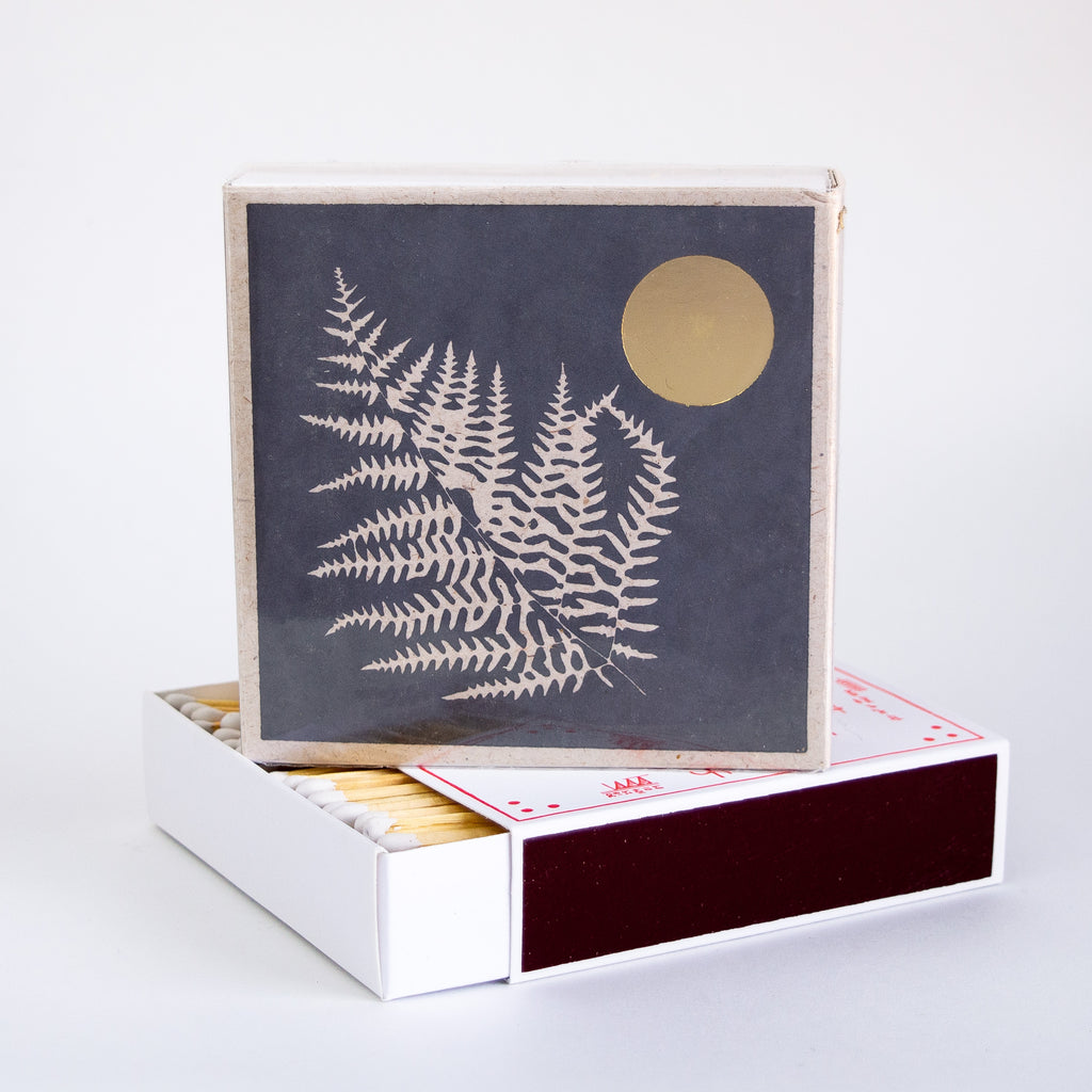 A box of matches that has a print of a white fern on a black backdrop with a gold moon.