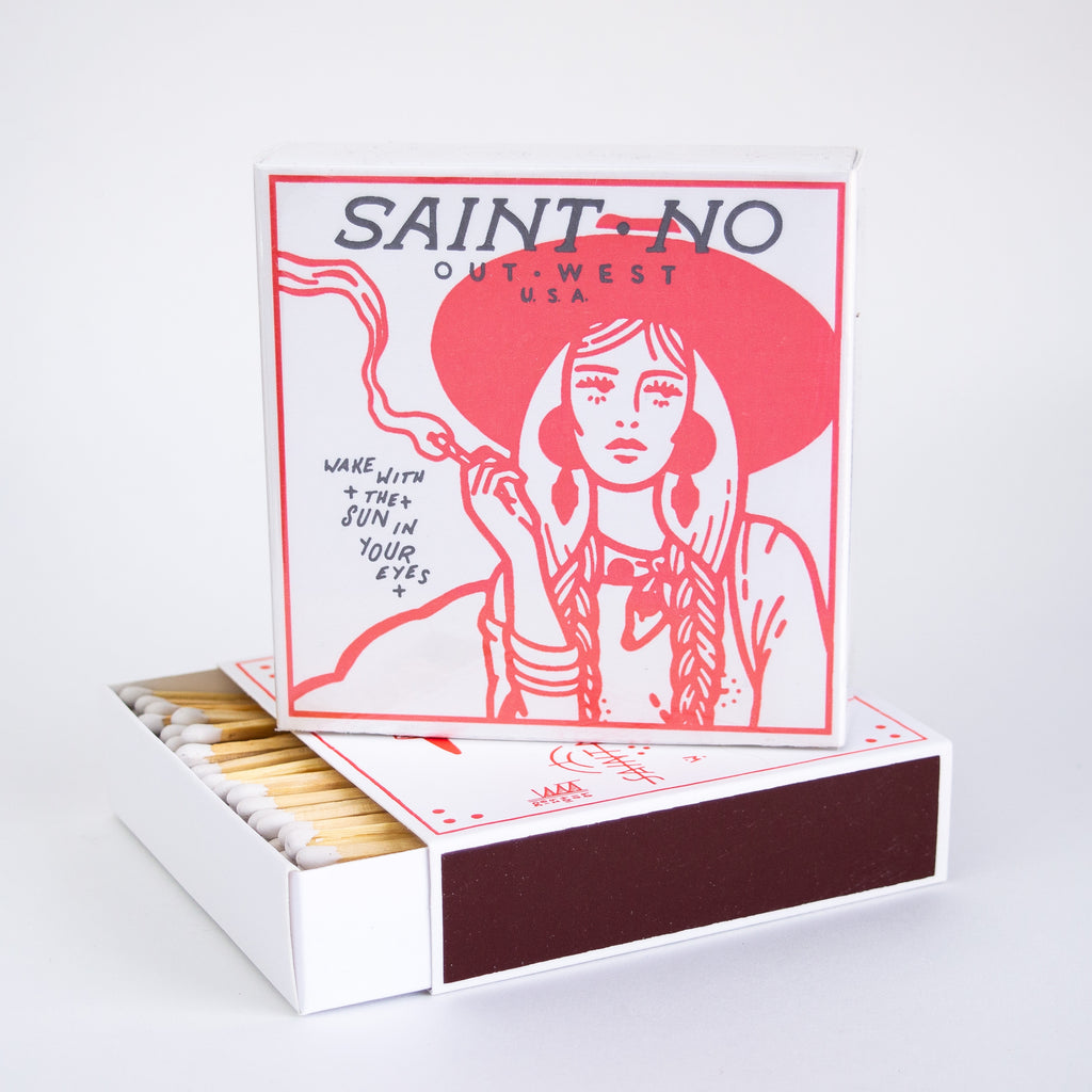 White square matchbox with a woman in southwestern garb holding a cigarette. There is black text on the box reading "Saint No Out West USA, Wake with the sun in your eyes." 