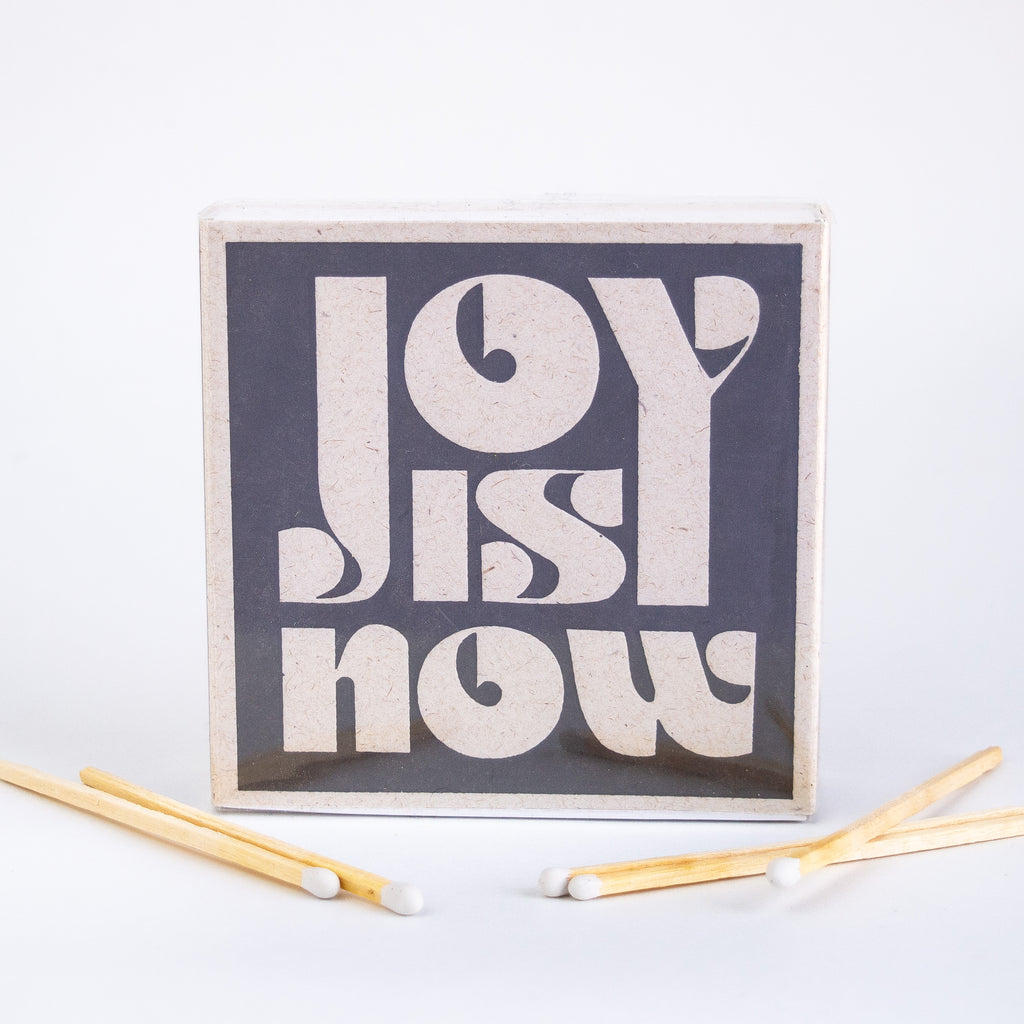 Square Matchbox with tan text reading "Joy is now."
